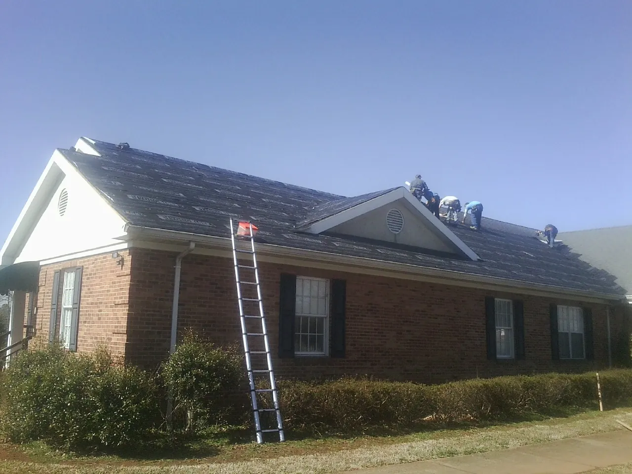 Crew tearing shingles off a brick ranch roof with a ladder set — Emergency Roof Repair work by our crew in Little Compton
