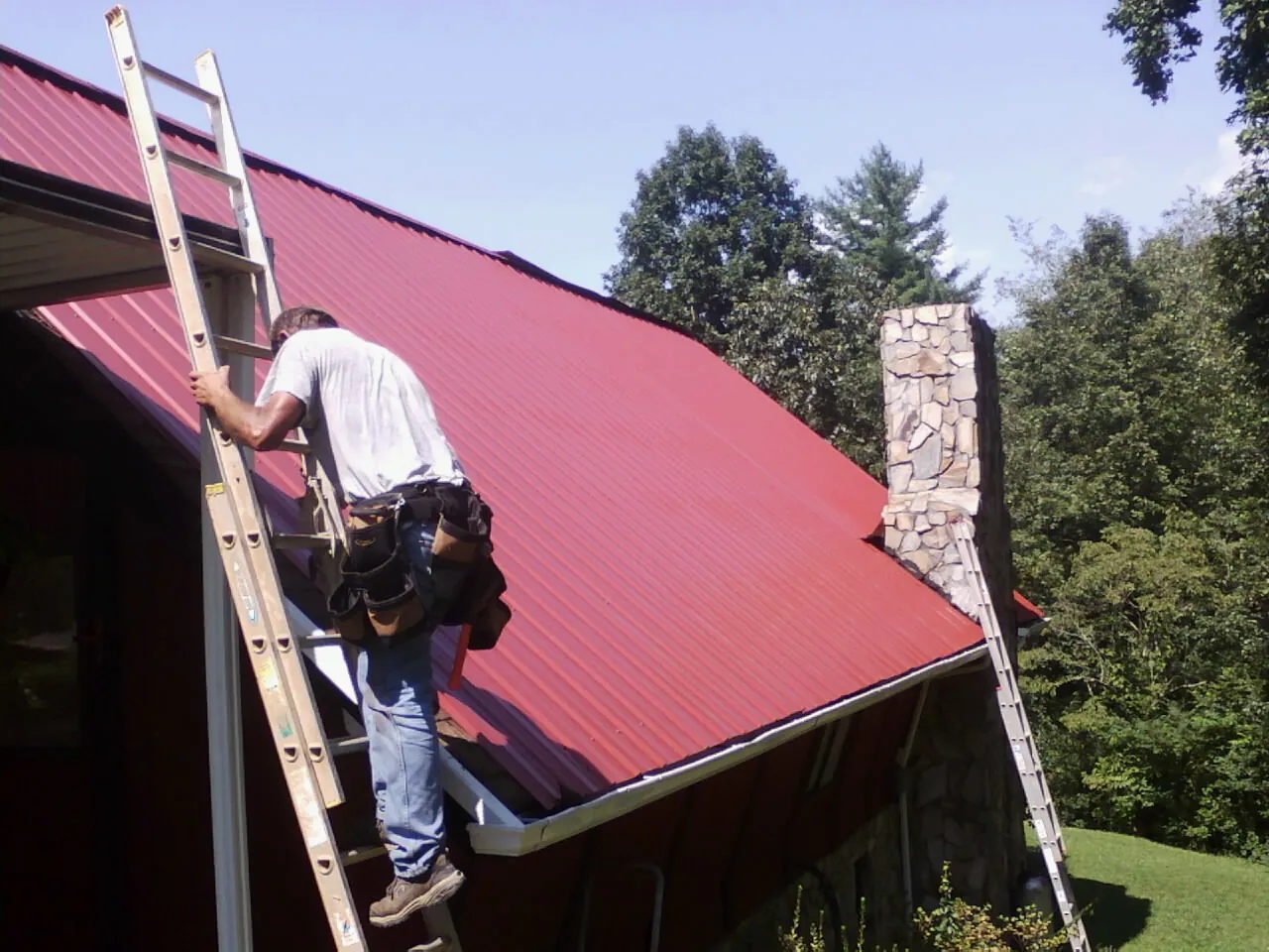 Roofer on a ladder beside a red metal roof and stone chimney — Soffit & Fascia Repair by Roofing Brothers in Little Compton