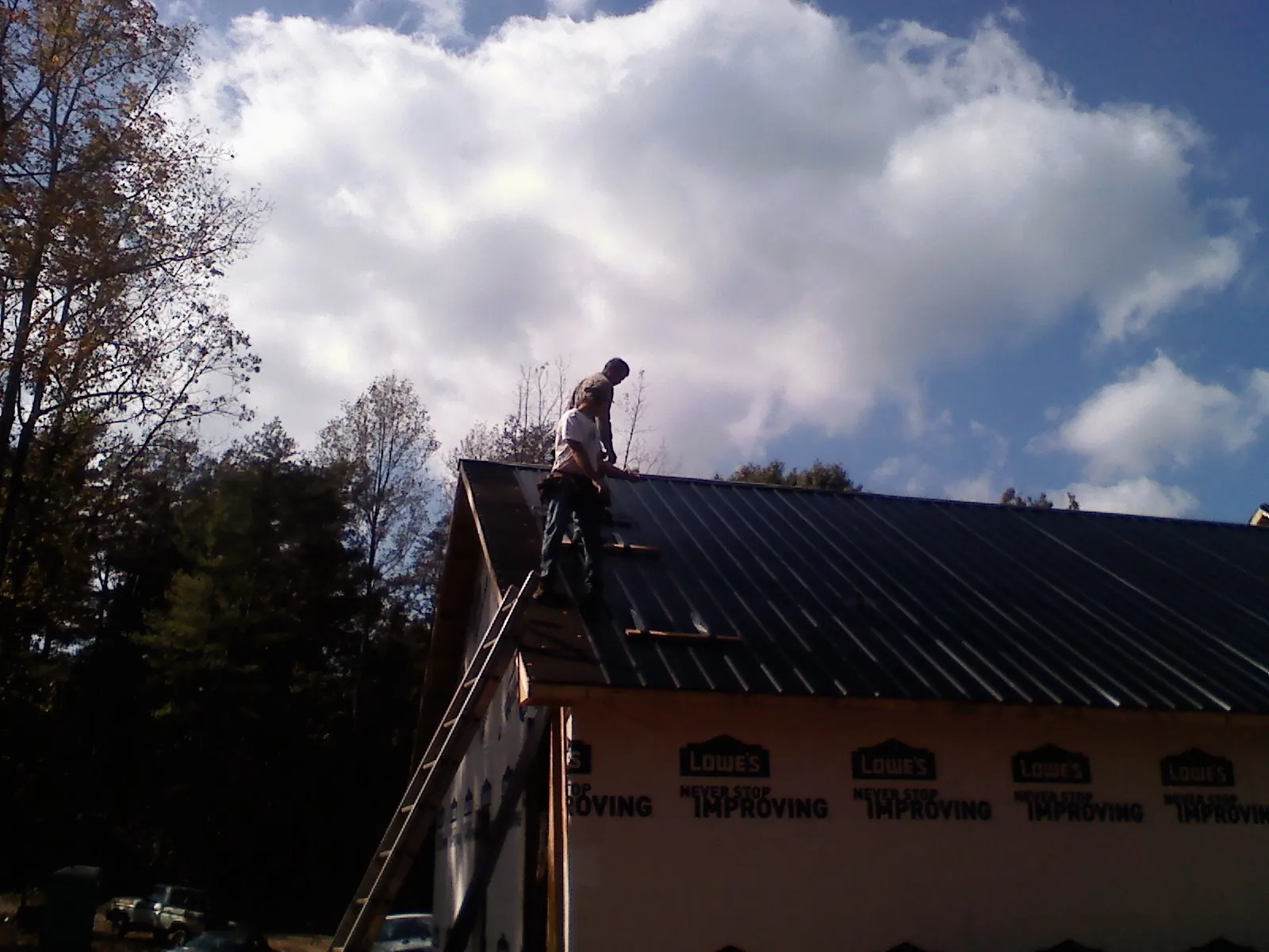 Roofer fastening dark metal panels high against a cloudy sky, from a Roof Inspection job in Little Compton